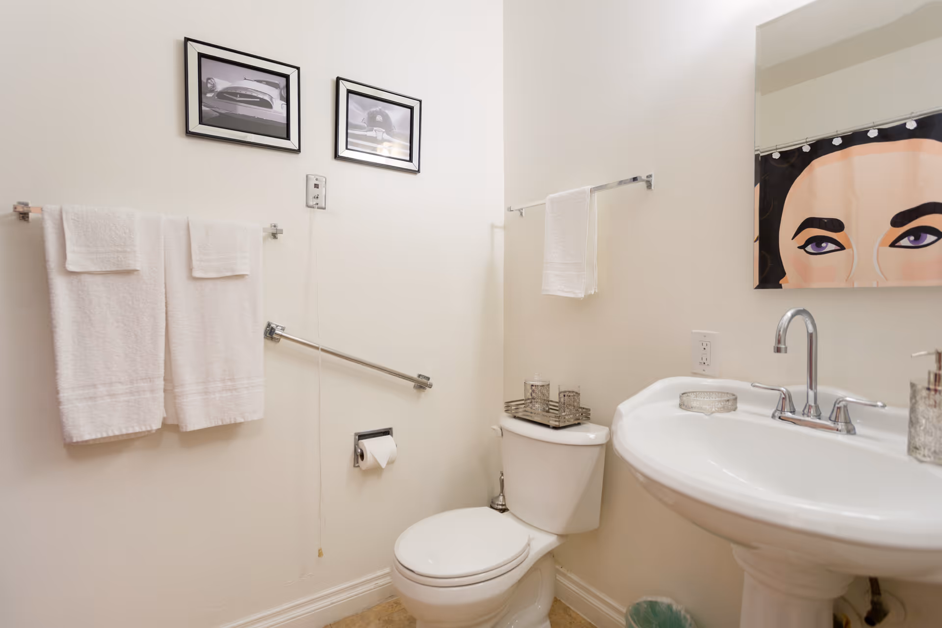 A clean bathroom with a white toilet and pedestal sink. Two white towels hang on a towel rack above a grab bar next to the toilet. A toilet paper holder is mounted on the wall. Above the sink is a mirror with a shower curtain reflected in it, showing a face with purple eyeshadow. Two framed black and white photos of cars are hung on the wall above the towels.
