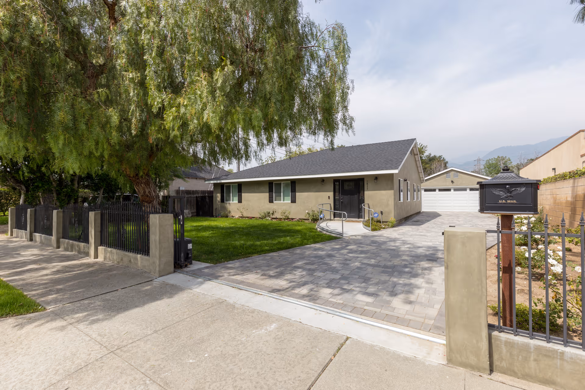 Exterior view of a single-story residential building with a gray roof and beige walls, a paved driveway leading to a detached garage, a green lawn, a large tree, and a black metal fence with a U.S. Mail mailbox at the entrance.