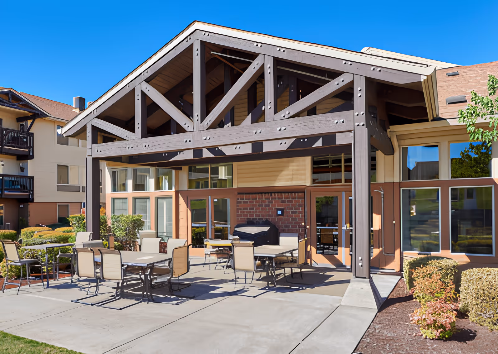 Outdoor patio area at Elliott Residence with several tables and chairs under a large wooden pergola attached to the building. The building has large windows and a brick wall section, with neatly trimmed bushes and landscaping around the patio. The sky is clear and blue.