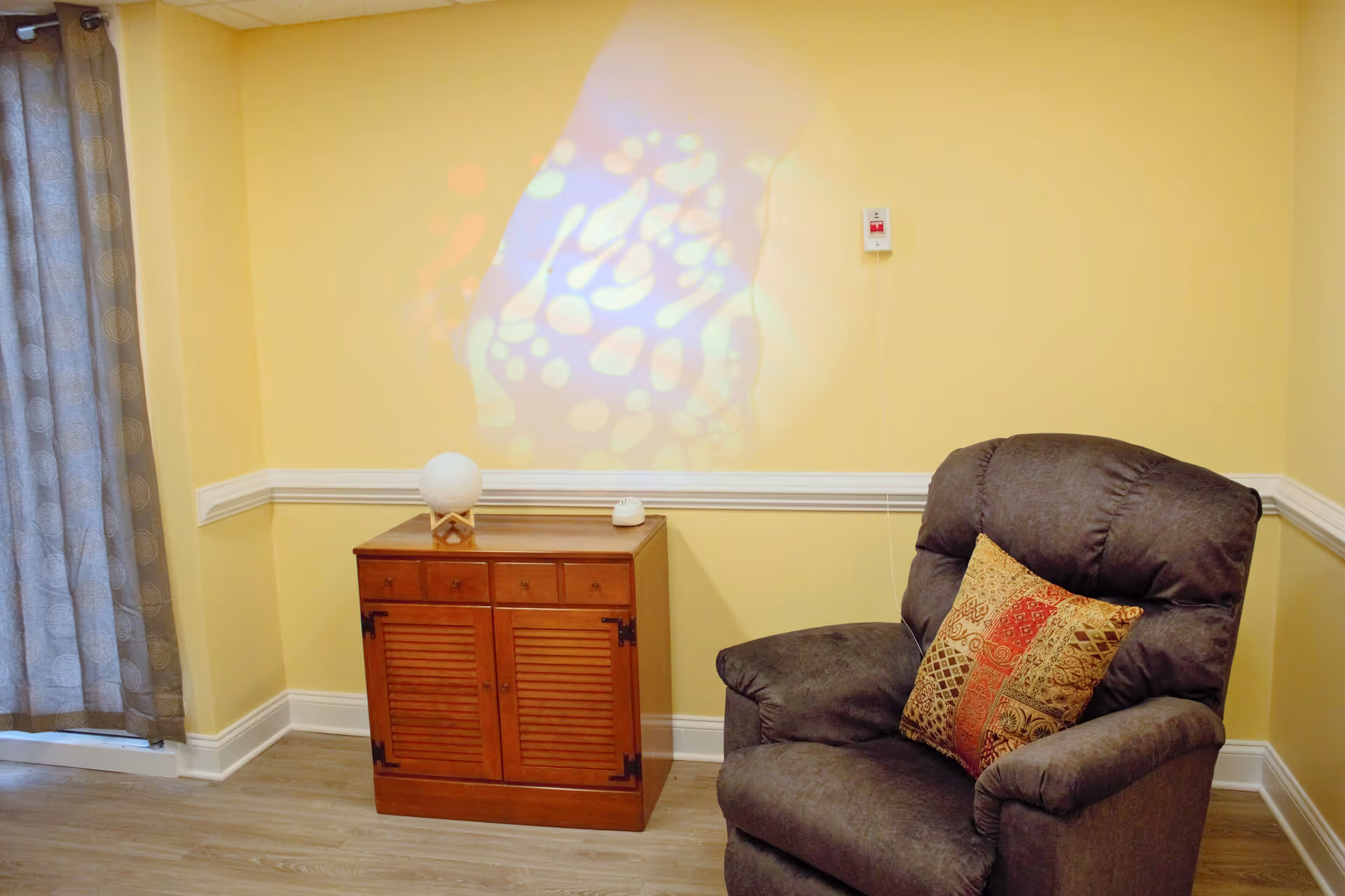 A cozy corner of a room with a dark gray recliner chair featuring a decorative patterned pillow. Next to the chair is a wooden cabinet with a spherical lamp and a small device on top. The walls are painted yellow with white trim, and a colorful light pattern is projected onto the wall above the cabinet. A curtain with a circular pattern hangs to the left.