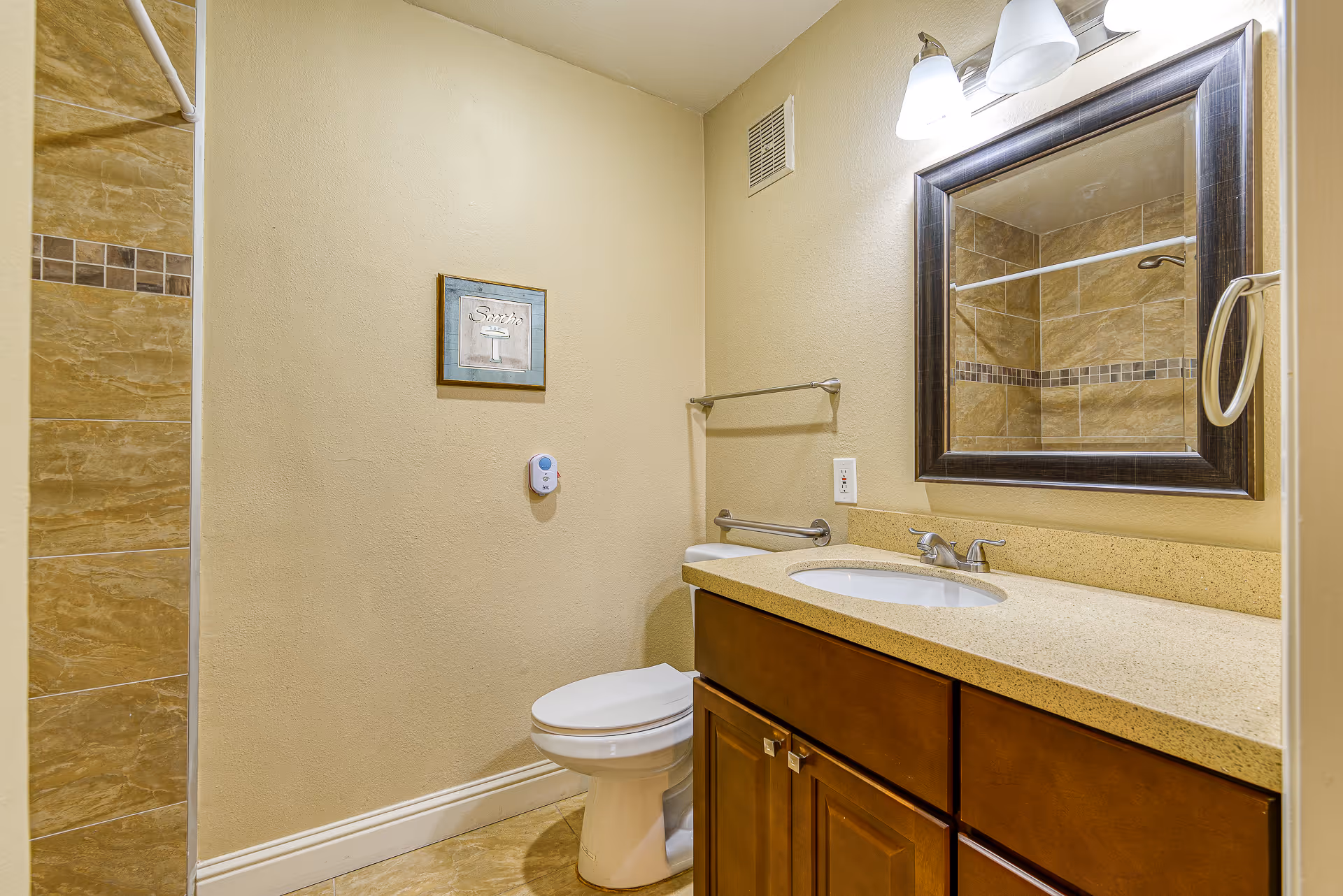 A bathroom with beige walls and tiled floor. It features a white toilet, a wooden vanity with a beige countertop and an oval sink, a large framed mirror above the sink, and a towel rack on the wall. There is a small framed picture on the wall and a shower with brown and beige tiles partially visible on the left side.