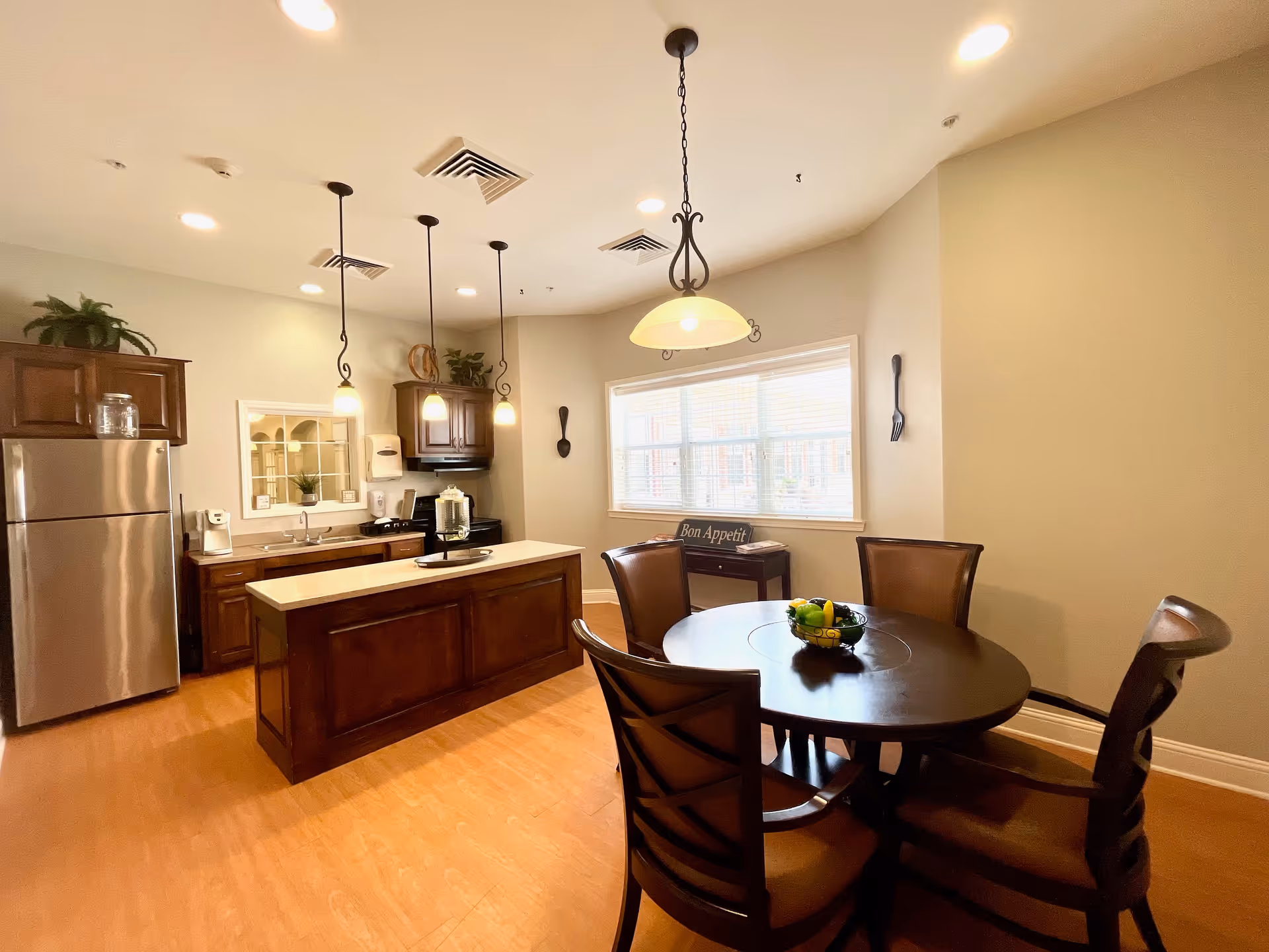 A warm, well-lit kitchen and dining area with wooden cabinets, a stainless steel refrigerator, a kitchen island with a light countertop, and three pendant lights hanging from the ceiling. There is a round dining table with four chairs and a bowl of fruit in the center. A window with blinds lets in natural light, and a small table beneath the window holds a 'Bon Appetit' sign. The walls are painted a light beige color, and the floor has a wood-like finish.