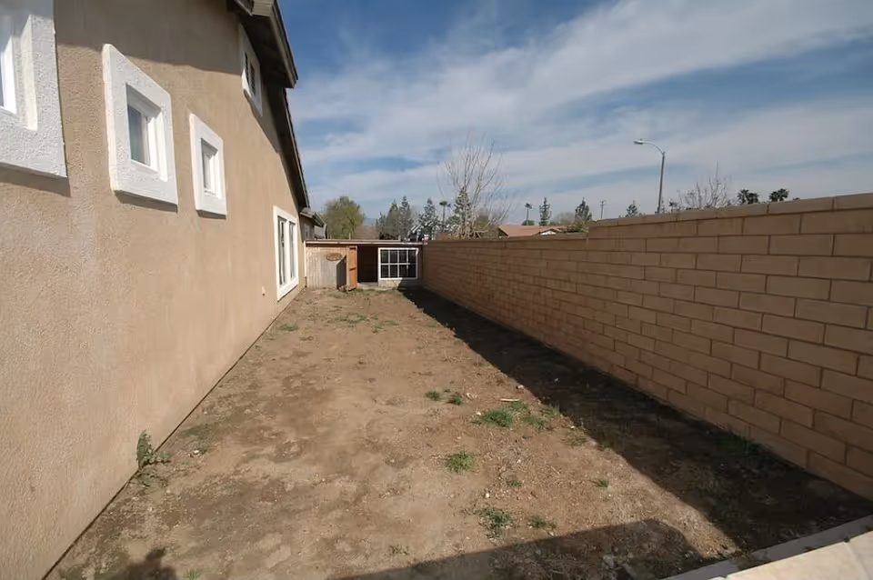 Narrow outdoor area beside a building with a dirt ground and a tall brick wall on the right side. The building has beige walls with white-framed windows. The sky is partly cloudy.