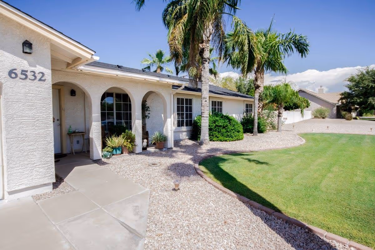 Front exterior of a single-story assisted living home with arched porch, palm trees, house number 6532, and a gravel and lawn yard.