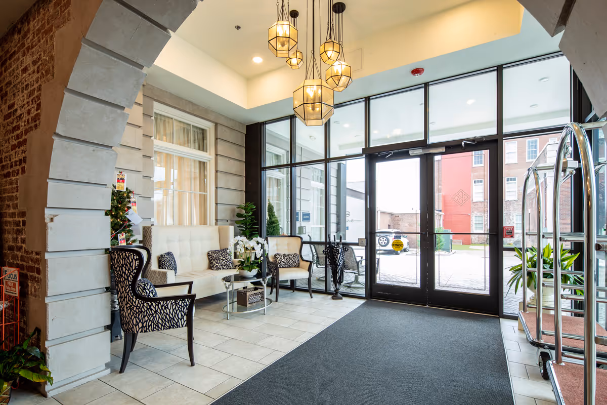 A bright and welcoming lobby area with large glass doors and windows allowing natural light to fill the space. The lobby features a seating area with a white cushioned bench, two patterned armchairs, a glass coffee table with a flower arrangement, and decorative plants. Modern pendant lights hang from the ceiling, and a luggage cart is visible on the right side near the entrance.