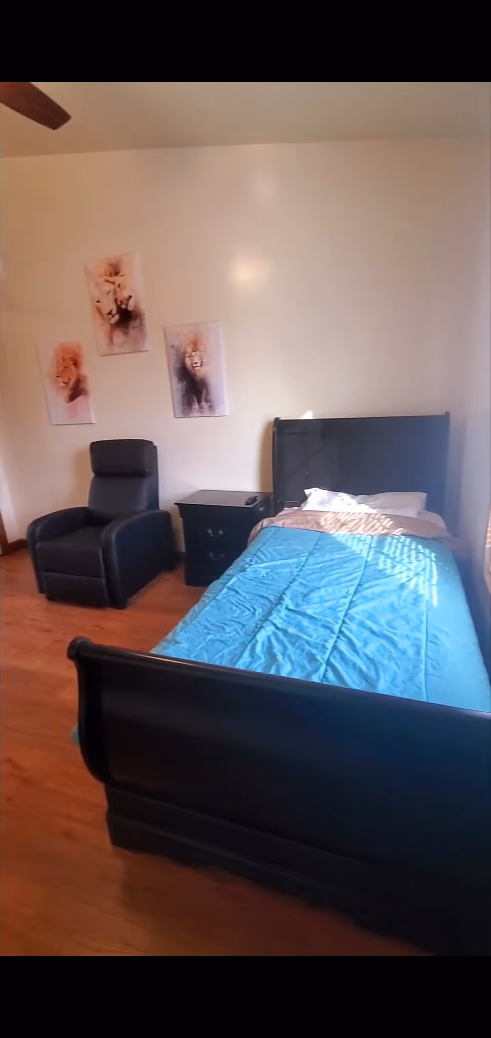 A bedroom with a single bed covered with a blue blanket and white pillow, a black nightstand with a remote control on it, a black recliner chair, and three animal-themed paintings on the wall. The room has wooden flooring and white walls.
