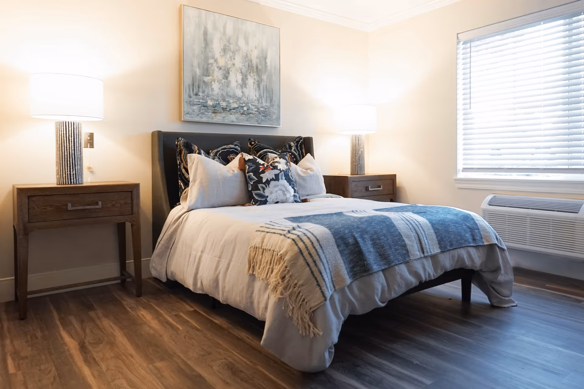 A cozy bedroom with a neatly made bed featuring multiple decorative pillows and a blue and white throw blanket. There are two wooden nightstands on either side of the bed, each with a lit table lamp. A large abstract painting hangs above the bed, and a window with white blinds is on the right side of the room. The floor is wooden, and the walls are painted a light beige color.