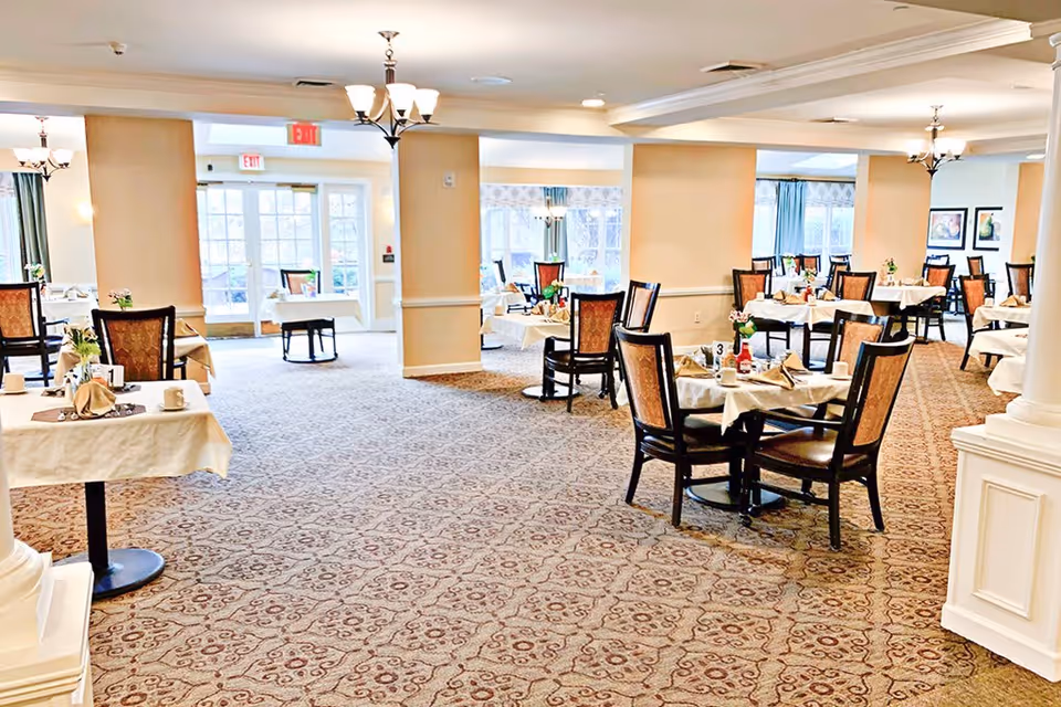 A spacious dining room with multiple round tables covered with white tablecloths, each set with napkins, cups, and small flower arrangements. The room has patterned carpet flooring, beige walls with white trim, and several chandeliers hanging from the ceiling. Large windows and glass doors allow natural light to fill the space.