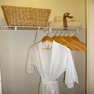 A white bathrobe hanging on wooden hangers in a closet with a woven basket and folded towels on the shelf.