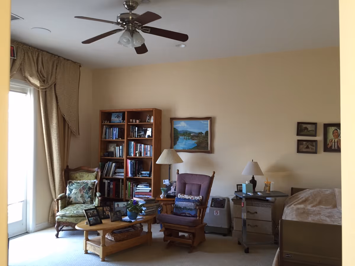 A cozy senior living room with beige walls and carpet. The room features a ceiling fan with lights, a wooden bookshelf filled with books and decorative items, a green upholstered armchair with a floral cushion, a wooden coffee table with framed photos and a potted plant, a purple cushioned rocking chair with a pillow, a side table with a lamp, a small medical device, and a hospital-style bed partially visible on the right. Several framed paintings and pictures hang on the walls.
