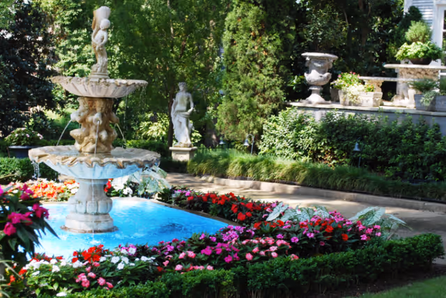 A beautifully landscaped garden area featuring a tiered stone fountain with water flowing, surrounded by vibrant pink, red, and white flowers and neatly trimmed green bushes. In the background, there are statues and large decorative urns placed on a raised stone platform, with lush green trees and shrubs providing a serene setting.