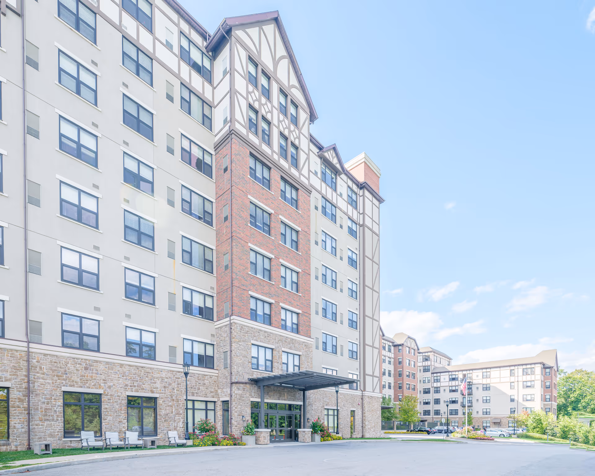 Front exterior of a multi-story senior living building with a covered entrance, brick and stucco facade, and adjacent wings under a blue sky.