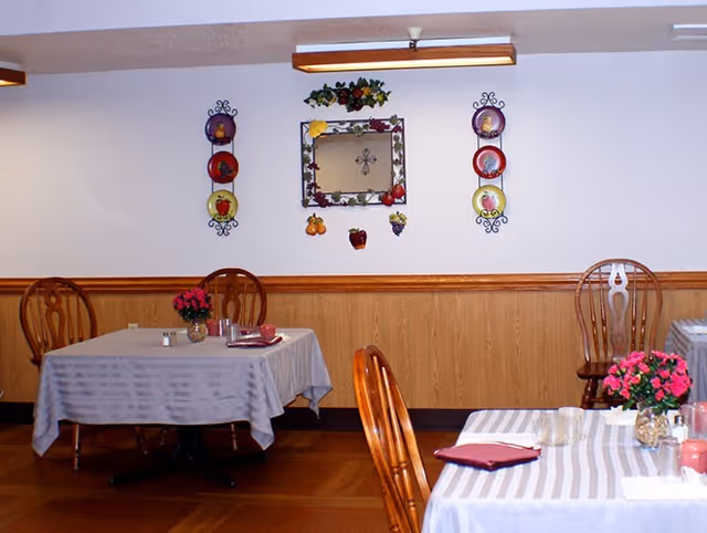 Interior view of a dining room with wooden chairs and tables covered with striped tablecloths. The tables have small flower arrangements and dining accessories. The wall features decorative plates and a mirror with fruit decorations around it.