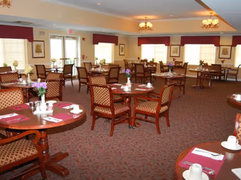A spacious dining room with multiple round wooden tables and cushioned chairs arranged neatly. Each table is set with white cups, saucers, napkins, and small flower vases. The room has large windows with red valances, beige walls, and carpeted flooring. Ceiling lights with chandeliers provide warm lighting.