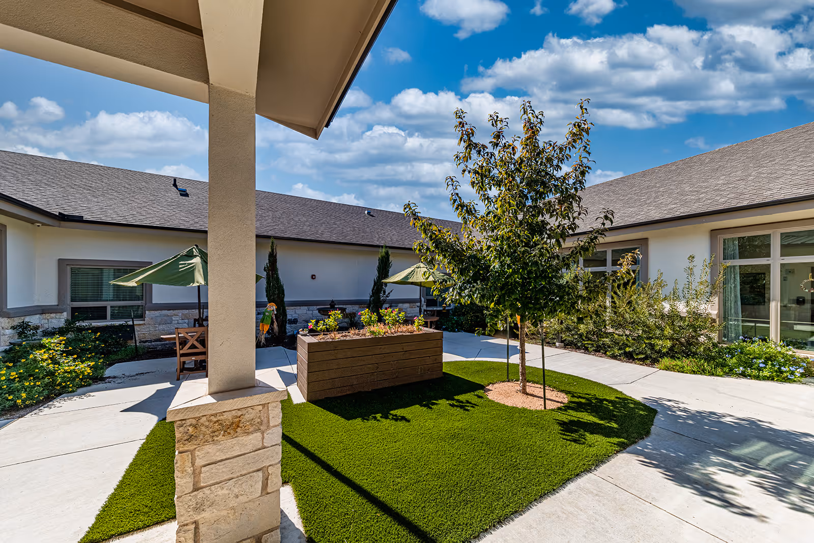 Outdoor courtyard area at Volante Senior Living of Sage Spring featuring a small tree planted in a circular patch of artificial grass, a raised wooden planter box with flowers, concrete walkways, patio tables with green umbrellas, and a building with large windows under a blue sky with scattered clouds.