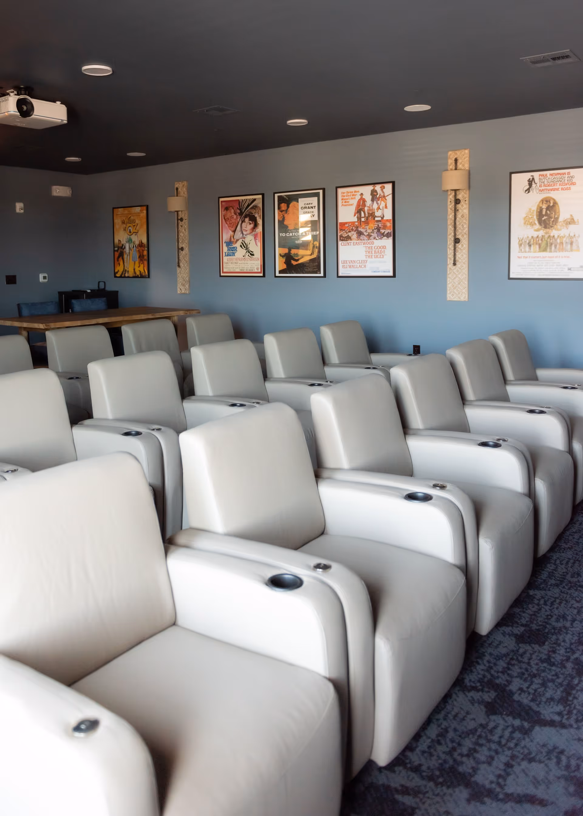 A room with multiple rows of white leather recliner chairs arranged facing a projector. The walls are painted blue and decorated with framed vintage movie posters. There is a wooden table with blue chairs in the back corner of the room.