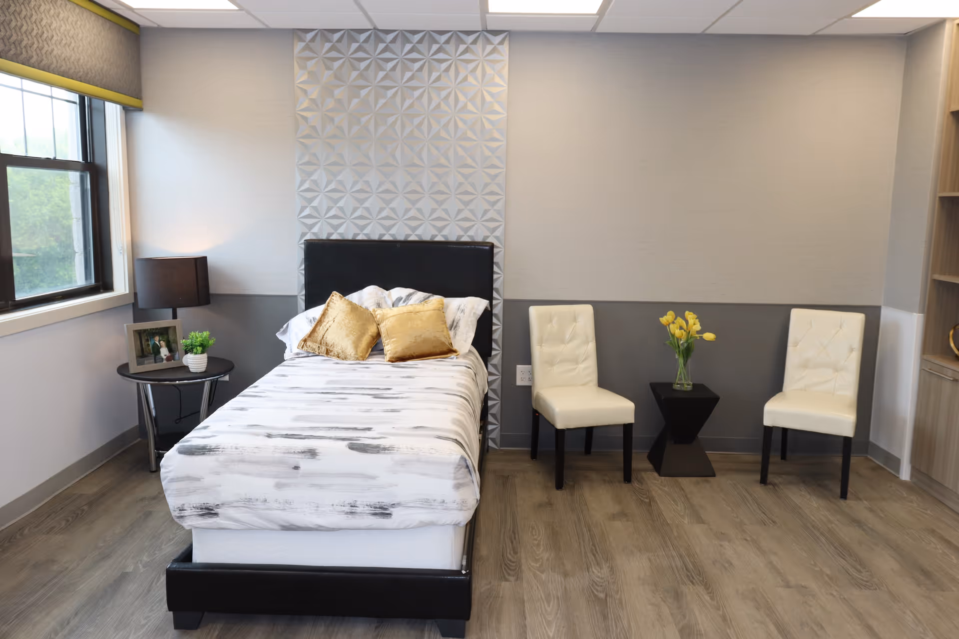 A neatly made single bed with white and gray bedding and two gold pillows in a modern room. To the left of the bed is a small round table with a black lamp, a framed photo, and a small potted plant. On the right side of the room, there are two white cushioned chairs with a small black table between them holding a vase with yellow flowers. The room has light gray walls, a large window with a yellow valance, and wood flooring.