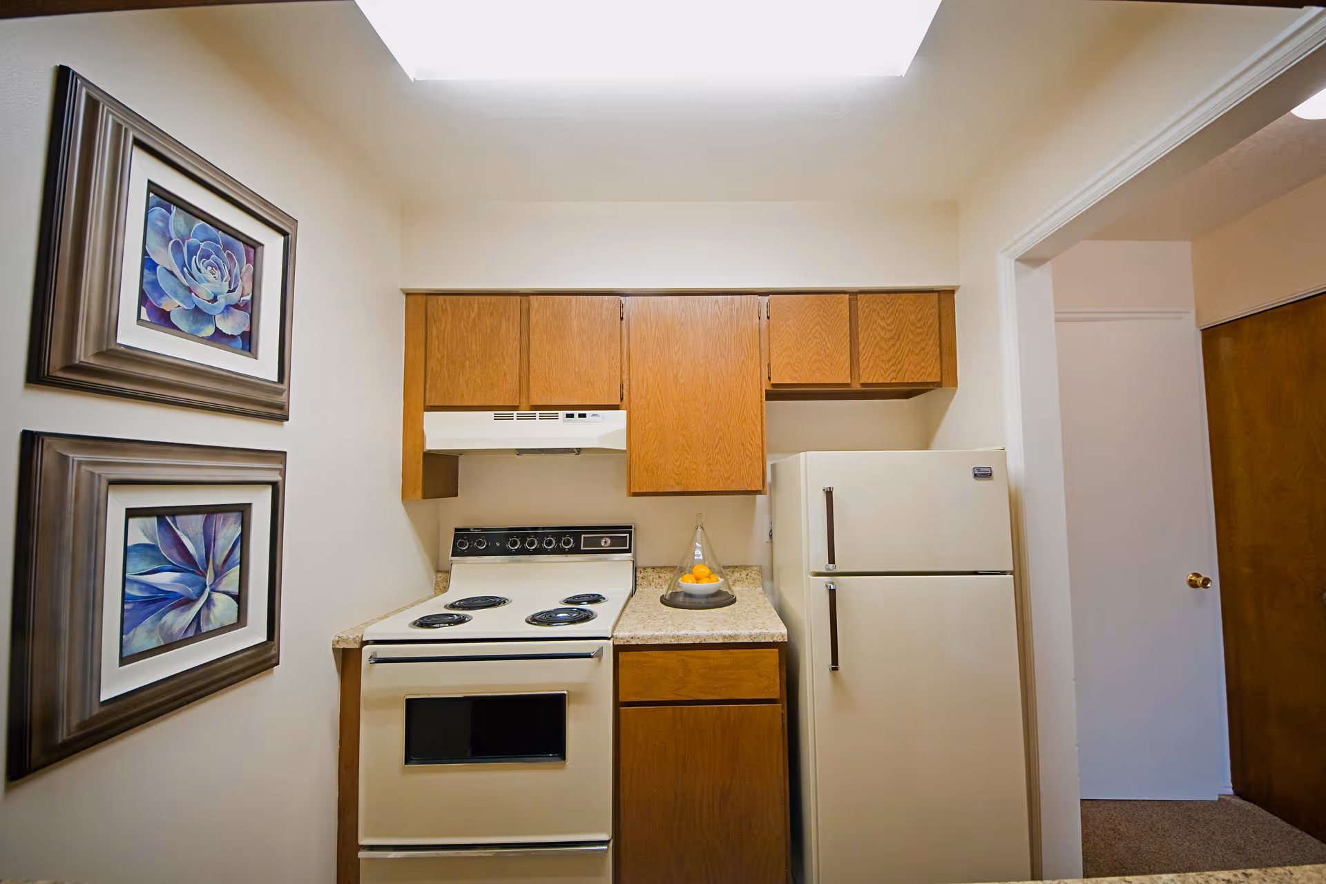 Small kitchen area with a white electric stove and oven, a white refrigerator, wooden cabinets above and below the countertop, and two framed floral paintings on the left wall.