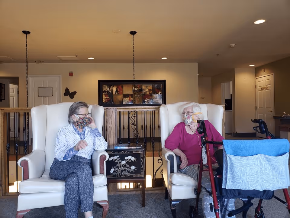 Two elderly women wearing face masks sitting in white armchairs in a well-lit common area. One woman is wearing a white patterned shirt and gray pants, while the other is wearing a magenta top and patterned pants with a red walker in front of her. The background shows a railing, a piece of wall art, and doors leading to other rooms.
