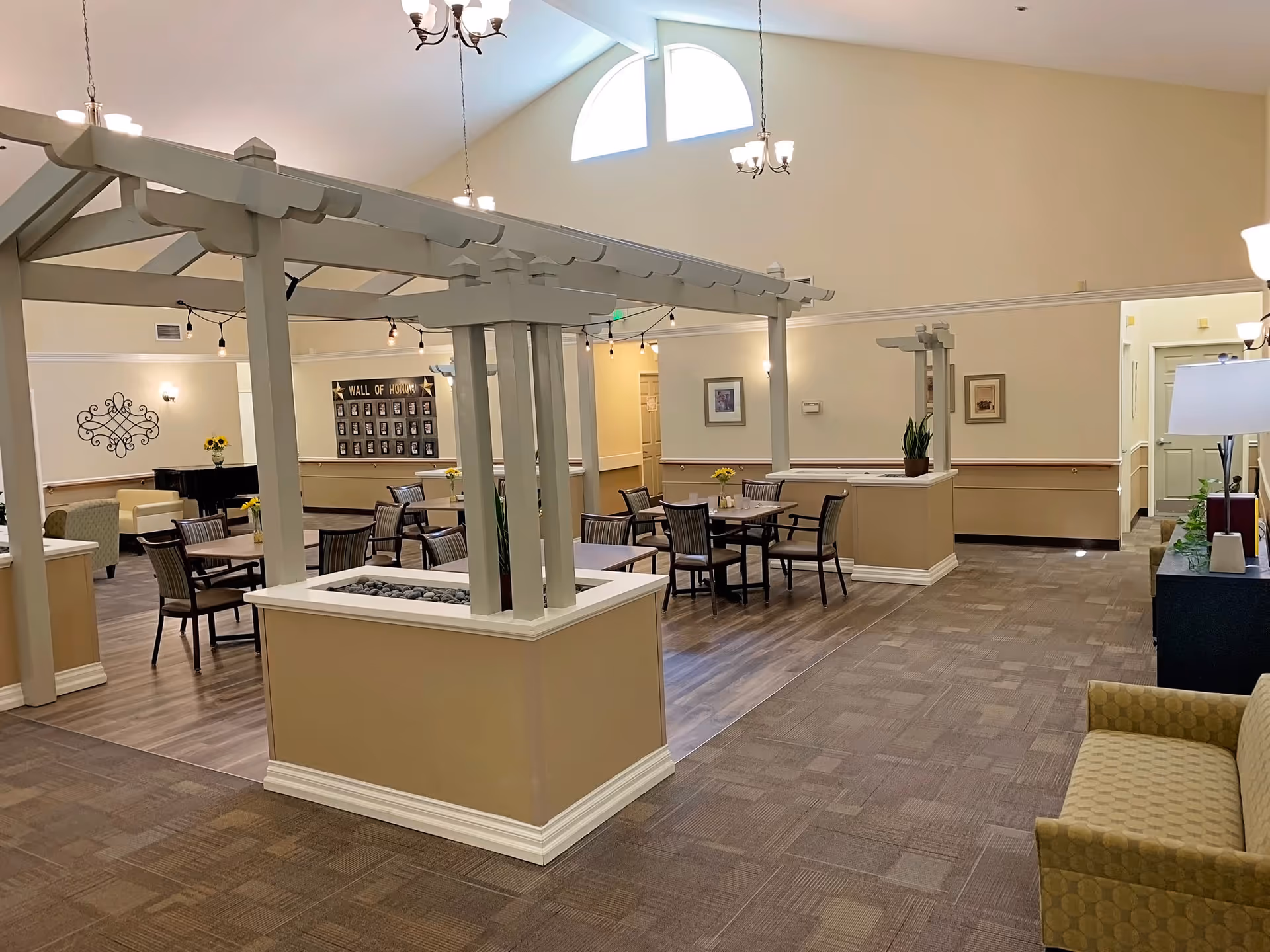 Interior view of a senior living facility common area with a high ceiling and large windows. The space features a pergola-like structure in the center, several tables and chairs arranged for dining or socializing, a piano in the background, and comfortable seating areas. The walls are decorated with framed pictures and a 'Wall of Honor' display.
