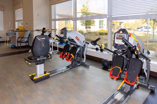A bright exercise room with large windows showing an outdoor view. The room contains two pieces of exercise equipment designed for seated workouts, with foot pedals and digital displays. There is also a chair, a table with a yellow exercise ball, and other small exercise accessories in the background.