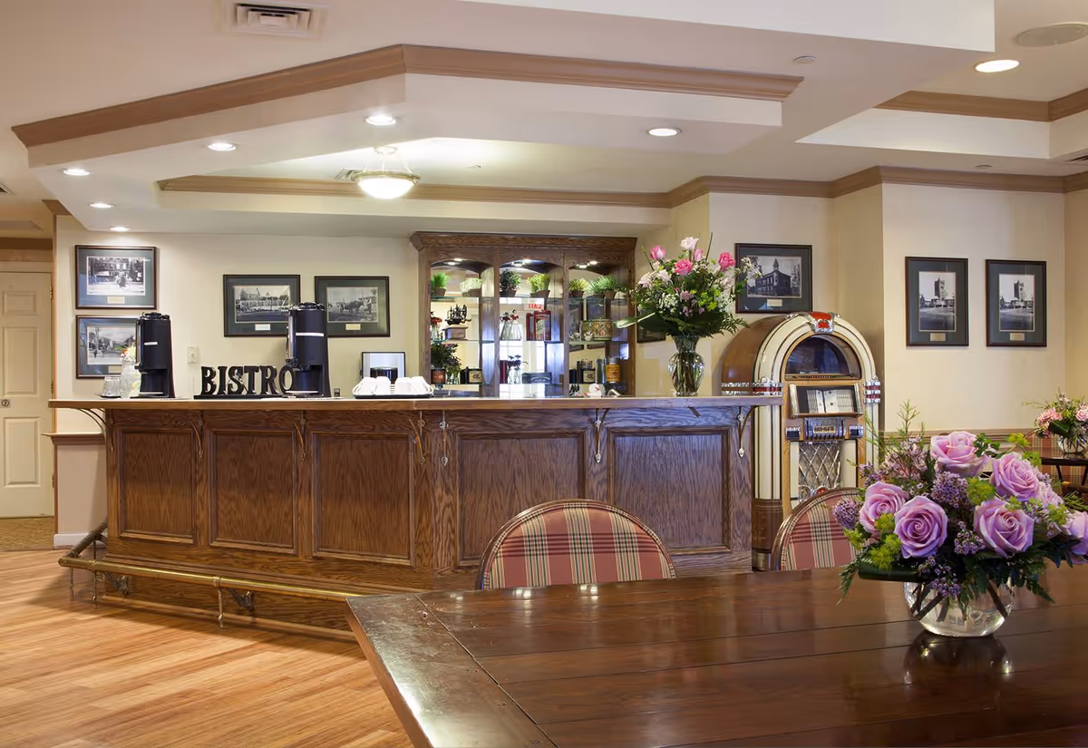 Interior view of a senior living facility's bistro area featuring a wooden counter with coffee dispensers and cups, a vintage jukebox, framed black and white photos on the walls, and floral arrangements on a wooden table with plaid upholstered chairs.