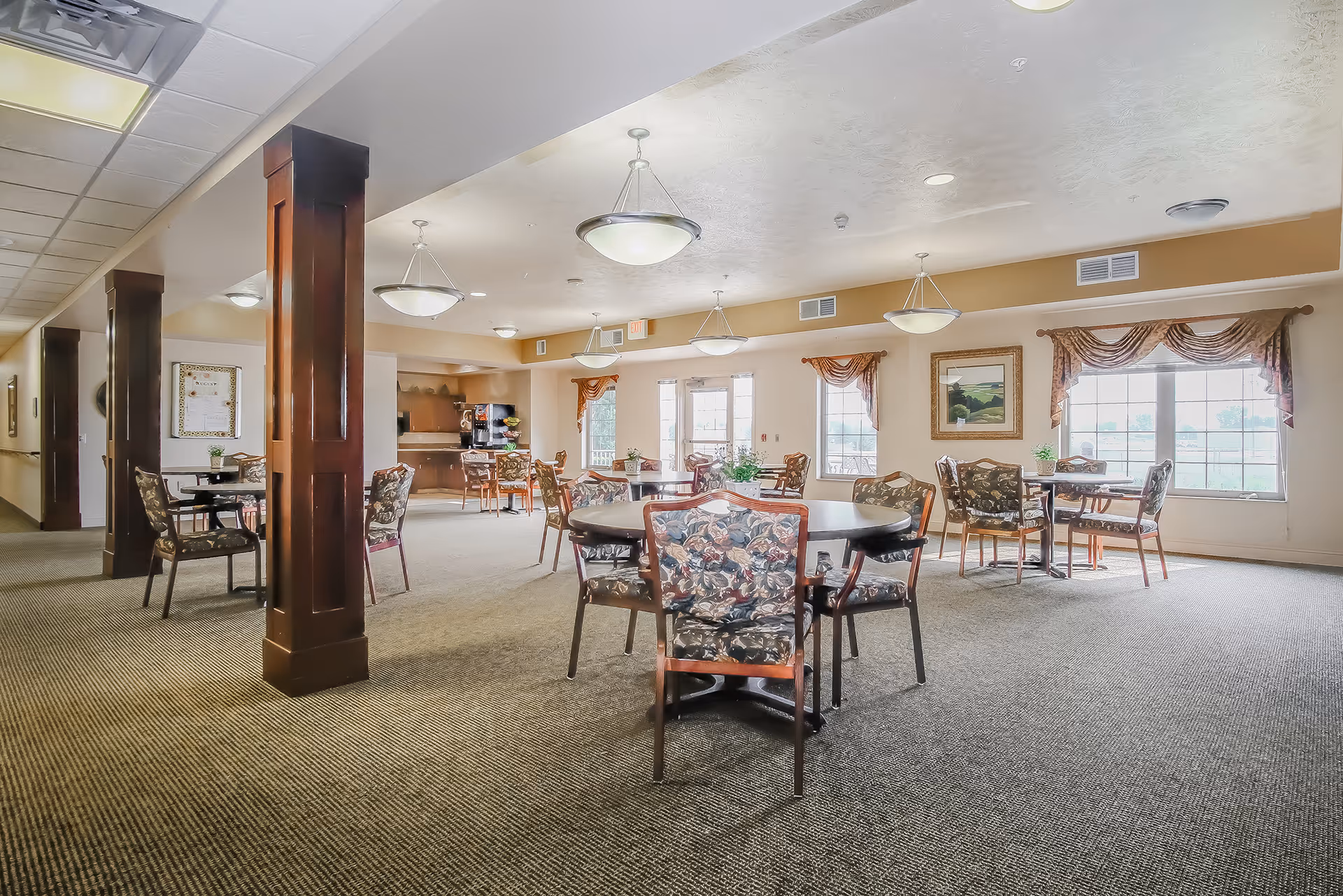 A spacious dining area in an assisted living facility with multiple round tables and floral upholstered chairs. The room is well-lit with hanging ceiling lights and natural light from large windows adorned with decorative curtains. There are wooden columns and a beverage station in the background.