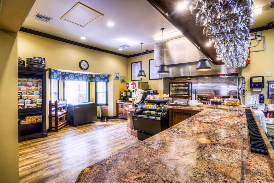 Interior view of a kitchen area in a senior living facility with a long granite countertop, hanging wine glasses above the counter, a display case with food items, coffee machines, and a window with a blue patterned valance letting in natural light.