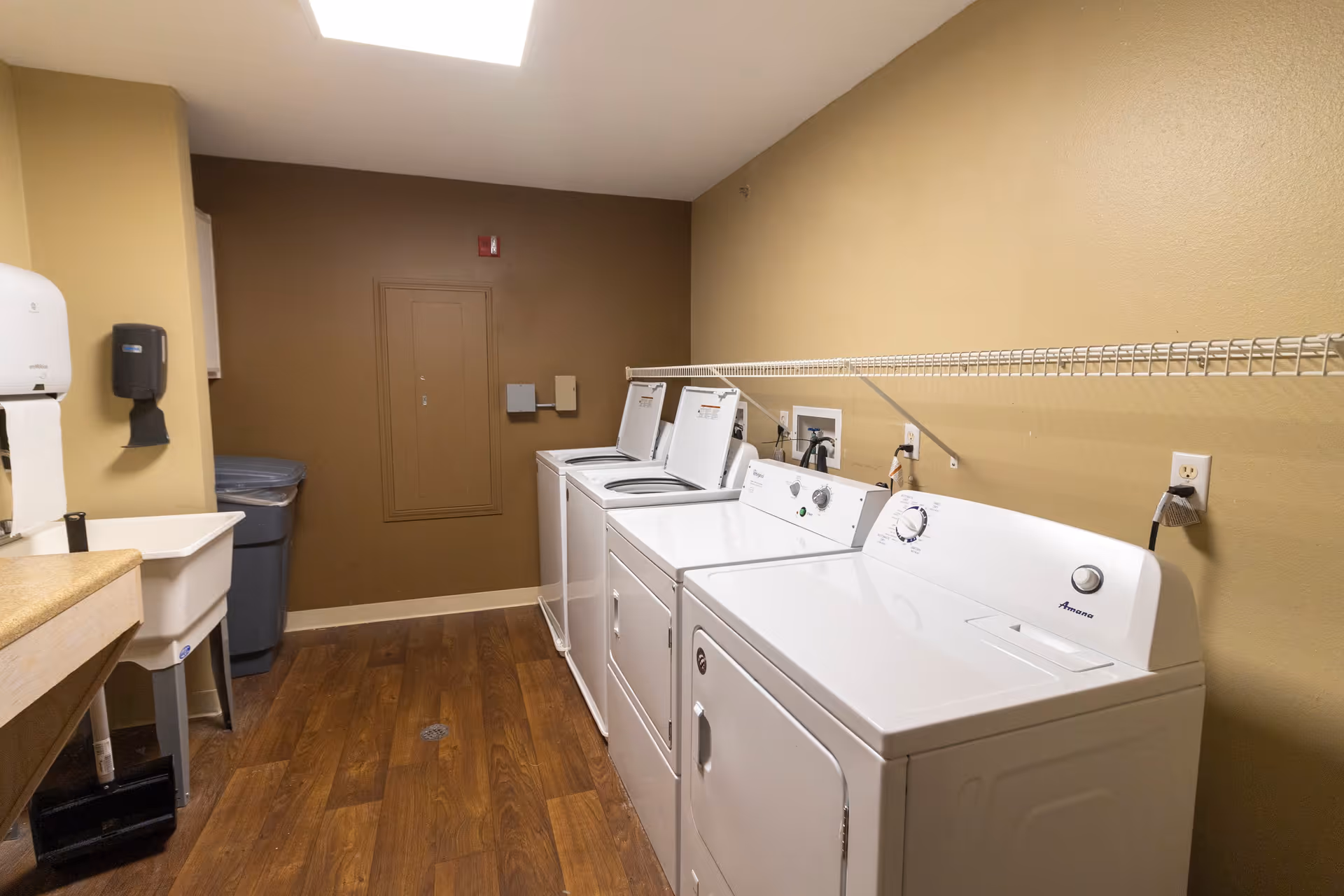 Laundry room with three white washing machines and dryers lined up against a beige wall. There is a white utility sink on the left side, a trash can, and a paper towel dispenser mounted on the wall. The floor is wood-patterned, and a wire shelf runs along the wall above the machines.