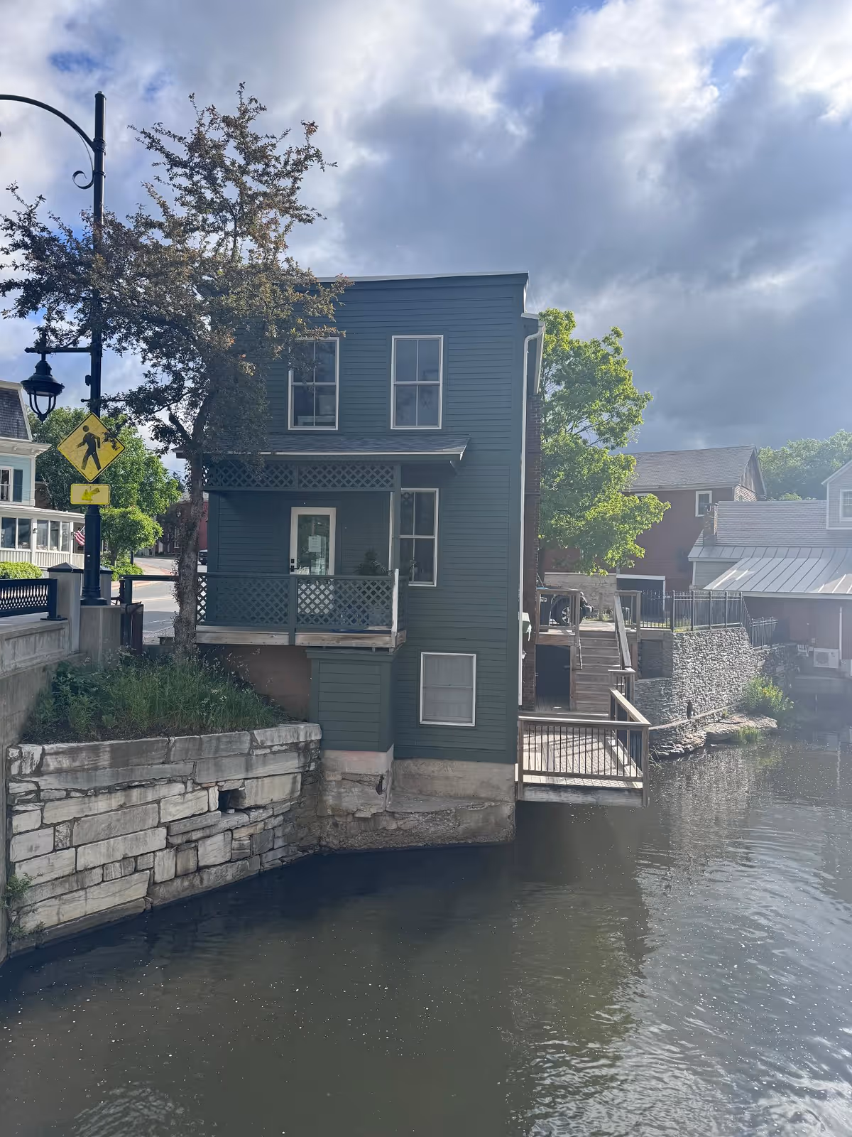 Two-story green wooden building with a small balcony overhanging a stone canal bank beside a river under a cloudy sky.