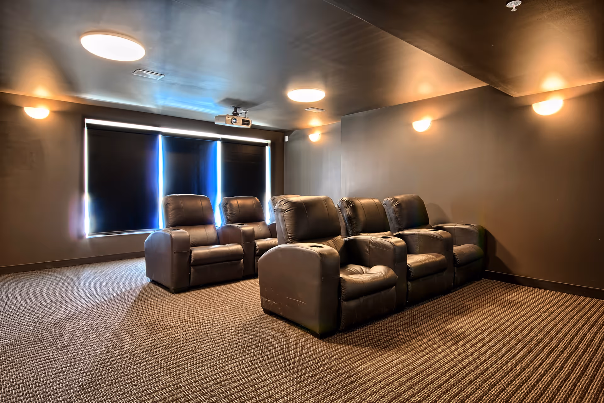 A small home theater room with six black leather recliner chairs arranged in two rows facing a window with black blackout curtains. The room has dark walls, carpeted floor, ceiling lights, and a projector mounted on the ceiling.