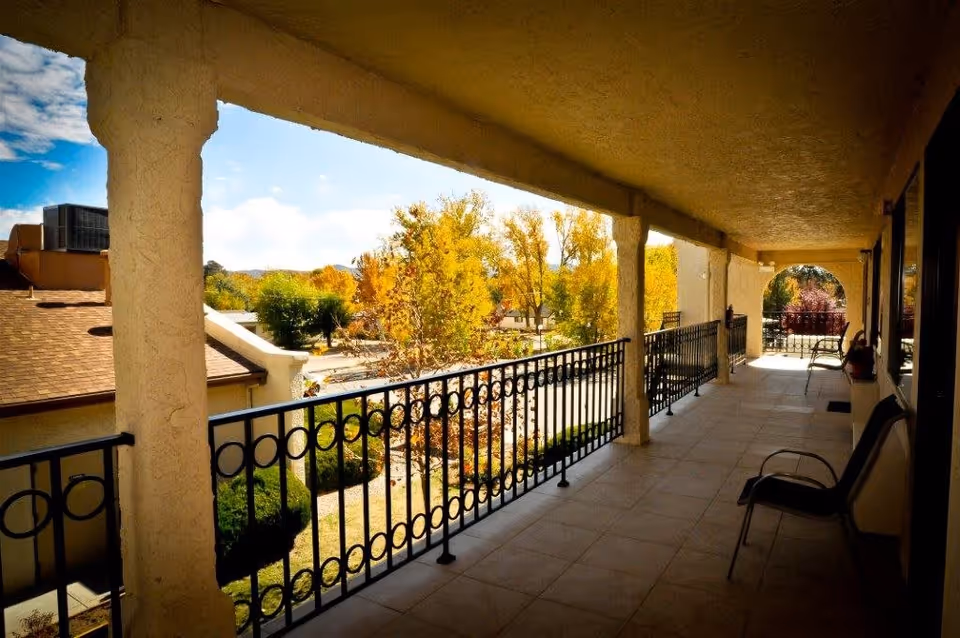 Covered outdoor balcony or walkway with tiled floor and black metal railing, overlooking trees with autumn foliage and a building with a brown roof. There are chairs placed along the right side near the windows.