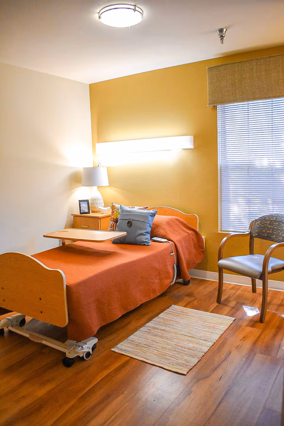 A nursing facility bedroom with a single hospital-style bed covered in an orange blanket and decorated with pillows. There is a small wooden nightstand with a lamp and a framed photo next to the bed. A wooden chair with armrests is positioned near a window with blinds. The room has wooden flooring and a small rug in front of the bed. The walls are painted in a combination of beige and mustard yellow.