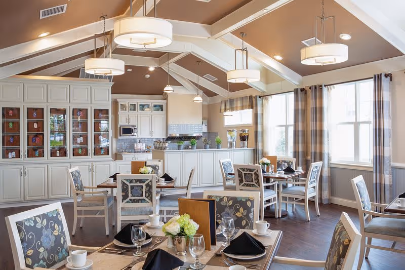 A bright and spacious dining room with multiple tables set for meals, featuring floral-patterned chairs, white cabinetry, large windows with plaid curtains, and modern pendant lighting hanging from a vaulted ceiling.