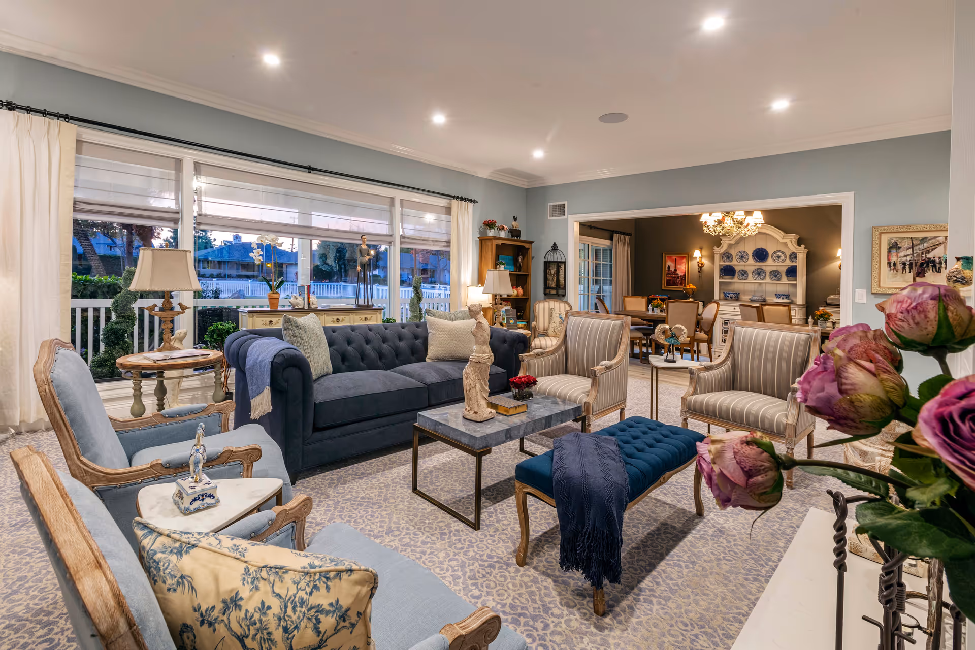 A cozy and elegant living room with a blue sofa, two striped armchairs, two light blue armchairs, a blue tufted bench, and a marble coffee table with decorative items. Large windows with white curtains let in natural light, and a dining area with a chandelier and a china cabinet is visible in the background. The room is decorated with flowers, lamps, and artwork.