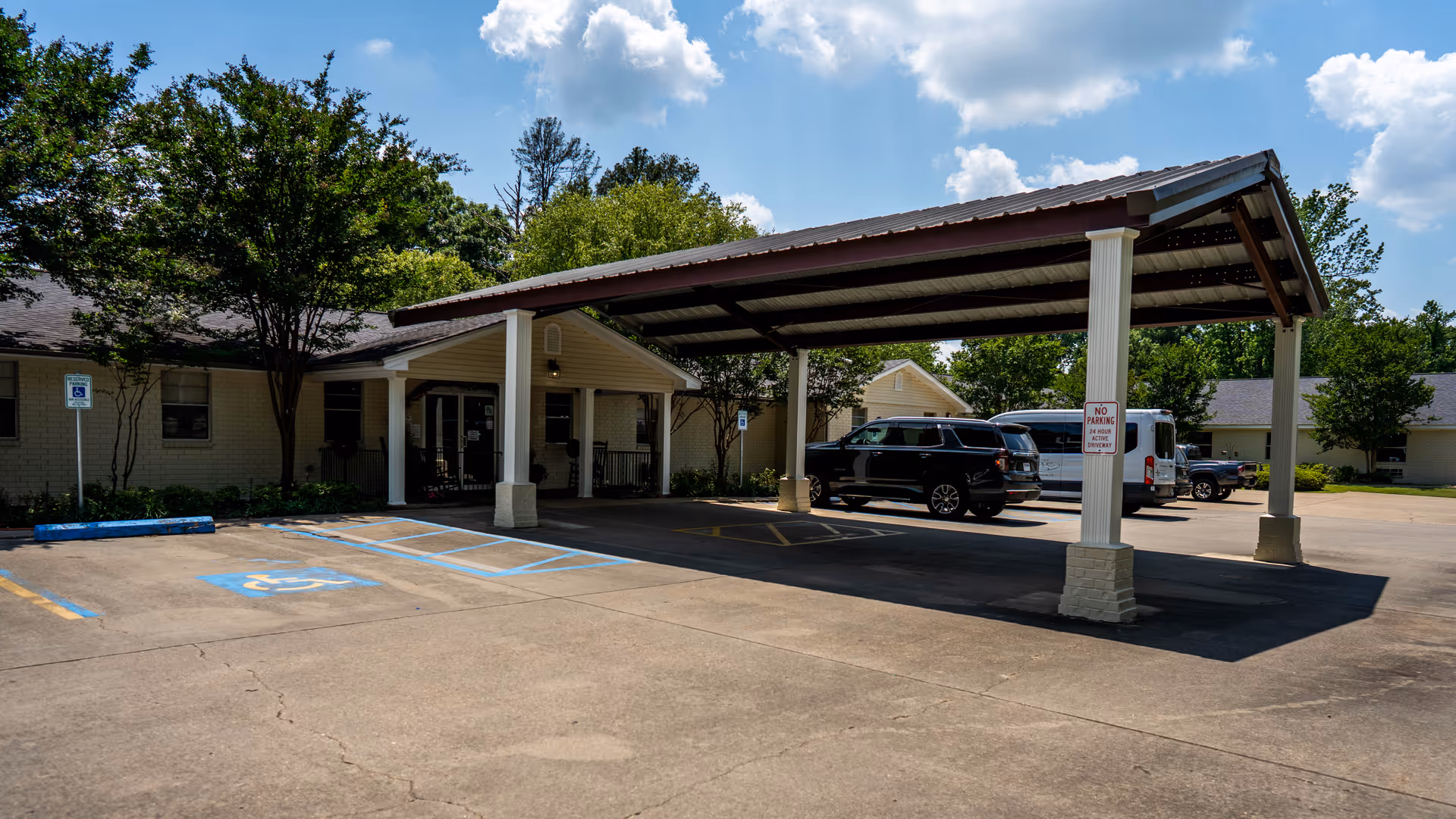 Exterior view of Lagniappe Health Care LLC showing a covered drop-off area with parked vehicles, a building entrance, and a handicapped parking space in front. Trees and a partly cloudy sky are visible in the background.