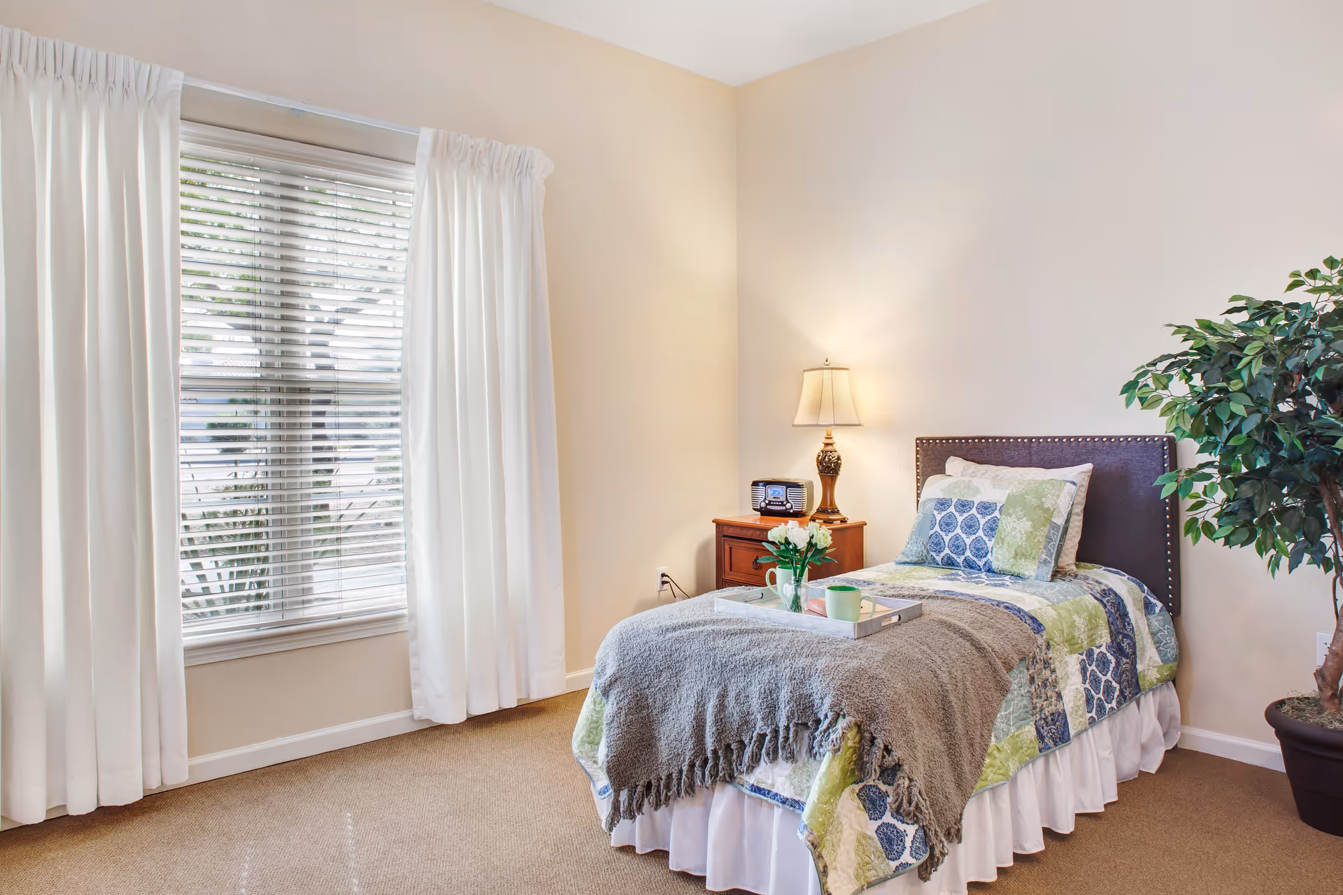 A cozy bedroom with a single bed covered in a patchwork quilt and a gray throw blanket. A tray with a green mug and a small vase of white flowers sits on the bed. Next to the bed is a wooden nightstand with a decorative lamp and a vintage-style radio. A large window with white curtains and blinds lets in natural light. A potted plant is placed in the corner of the room.