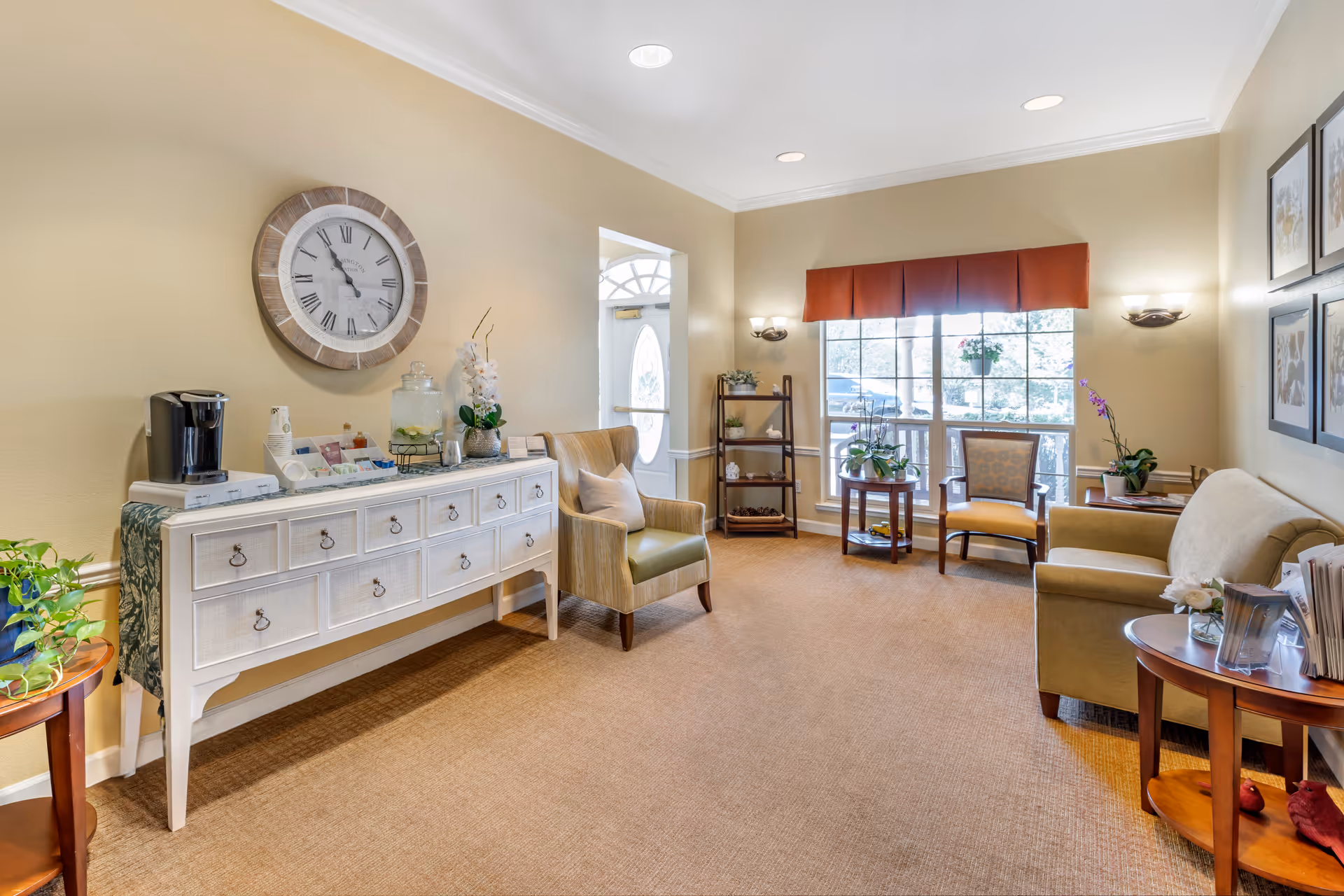 A bright and cozy living room area with beige walls and carpet. The room features a white sideboard with drawers, a large round wall clock above it, and a coffee maker on top. There are several chairs, including an armchair and a sofa, along with small tables holding plants and decorative items. A large window with a red valance lets in natural light, and framed artwork hangs on the wall.