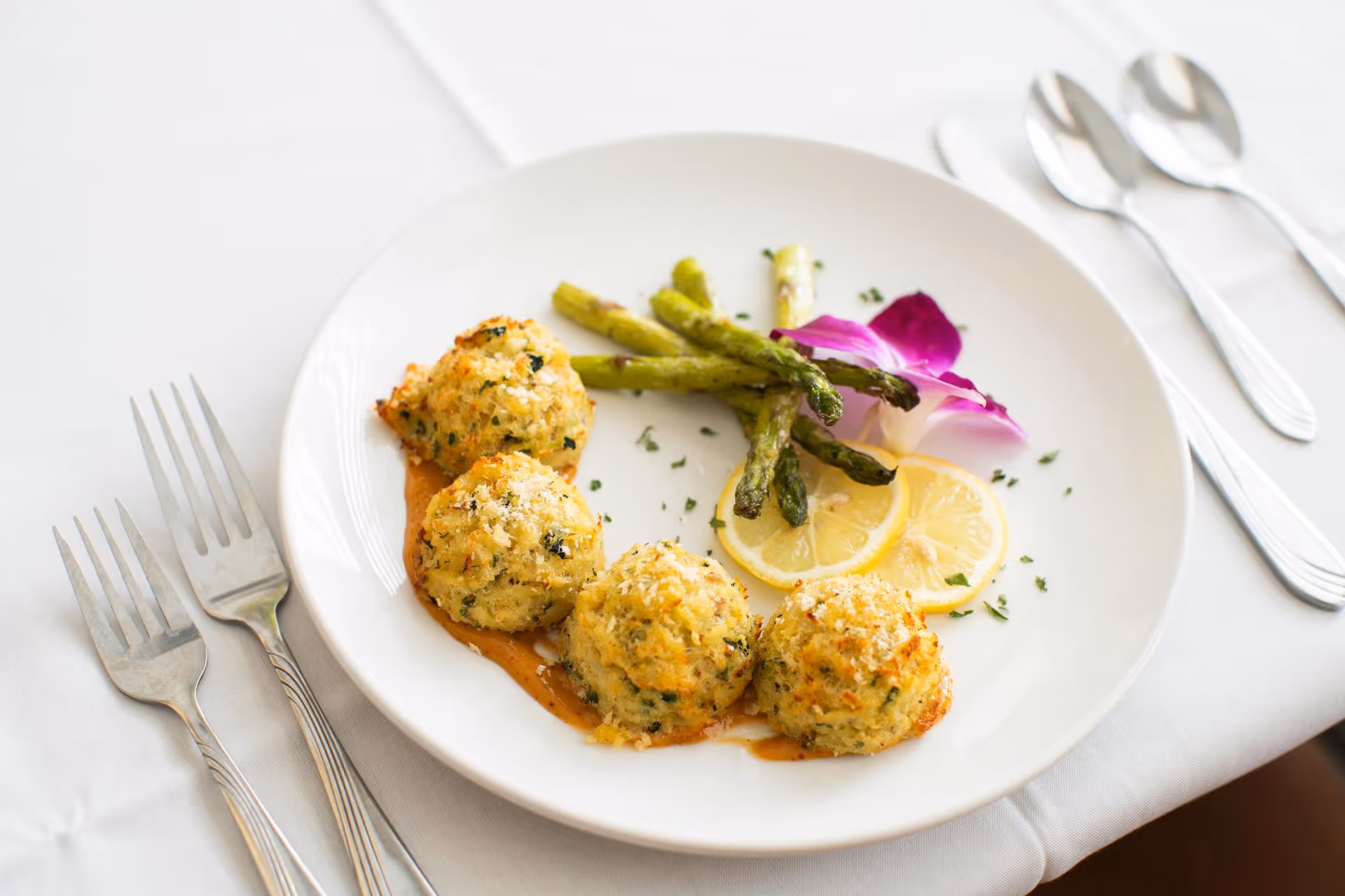 A white plate with four small golden baked rounds served with asparagus, lemon slices and an edible purple flower on a white tablecloth with forks and spoons.