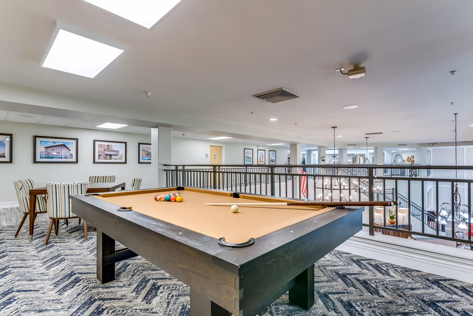 Interior view of a senior living facility's recreational area featuring a pool table with billiard balls and a cue stick on it. In the background, there is a seating area with a table and striped chairs, framed pictures on the wall, and a railing overlooking a lower level with chandeliers and an American flag.