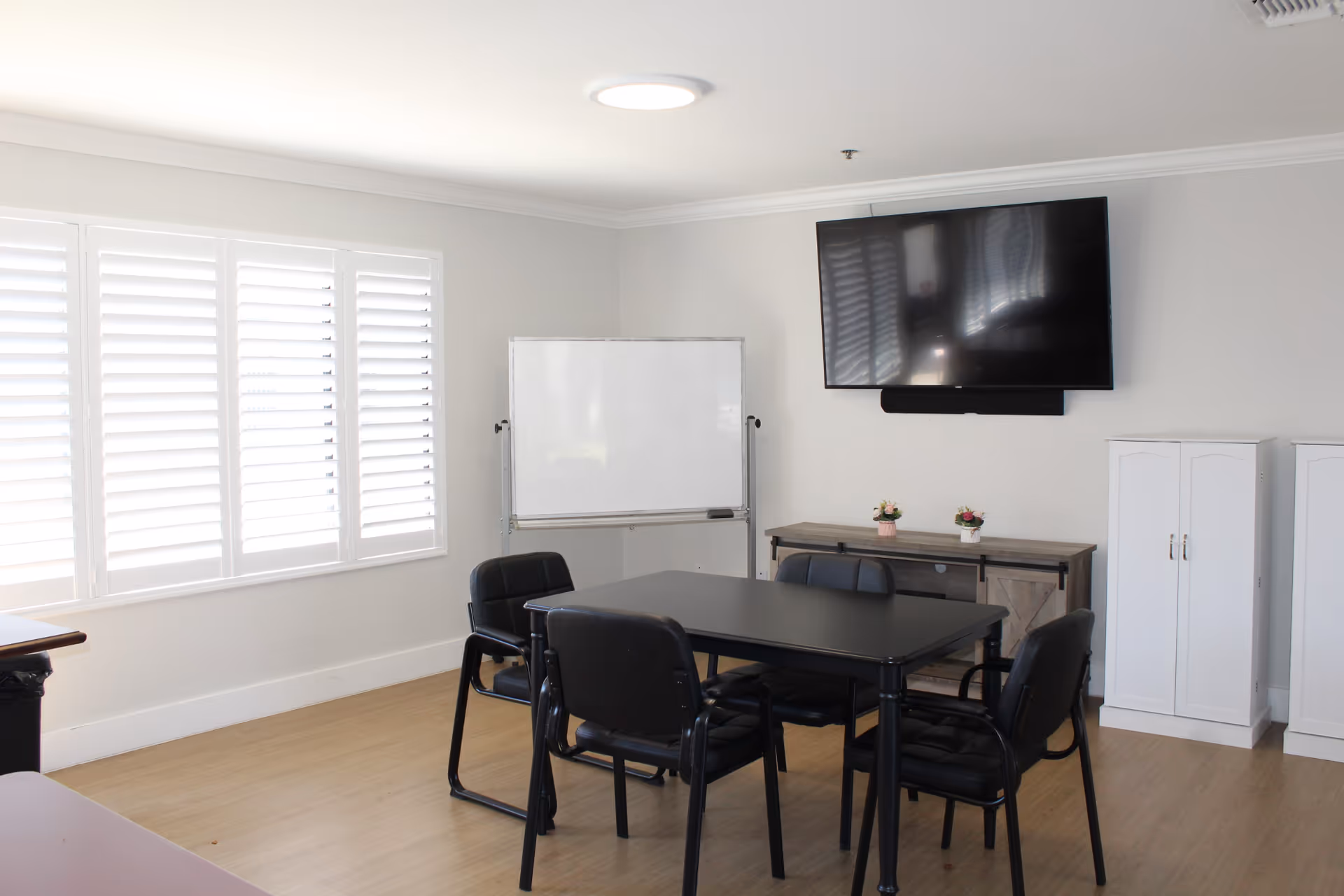 Small meeting/activity room with a table and chairs, a wall-mounted TV, whiteboard, and cabinets.