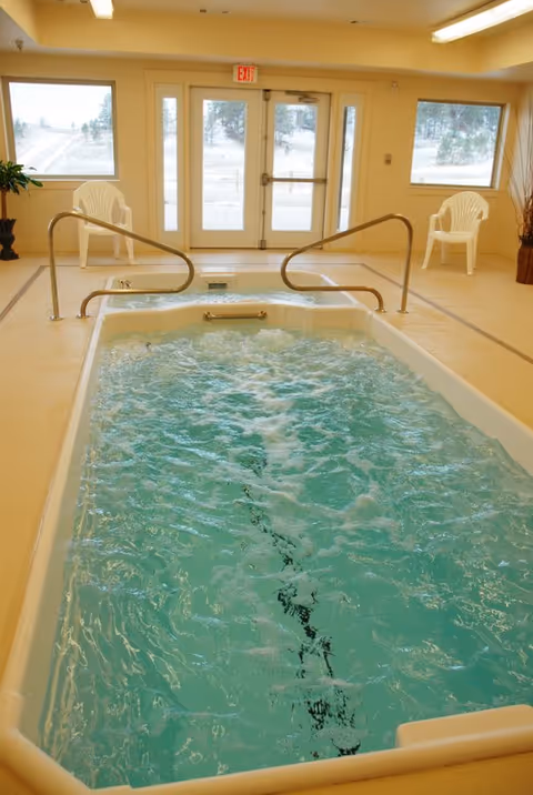 Indoor therapy pool with bubbling water and handrails on both sides, two white plastic chairs near large windows and glass doors showing an outdoor view with trees and snow.