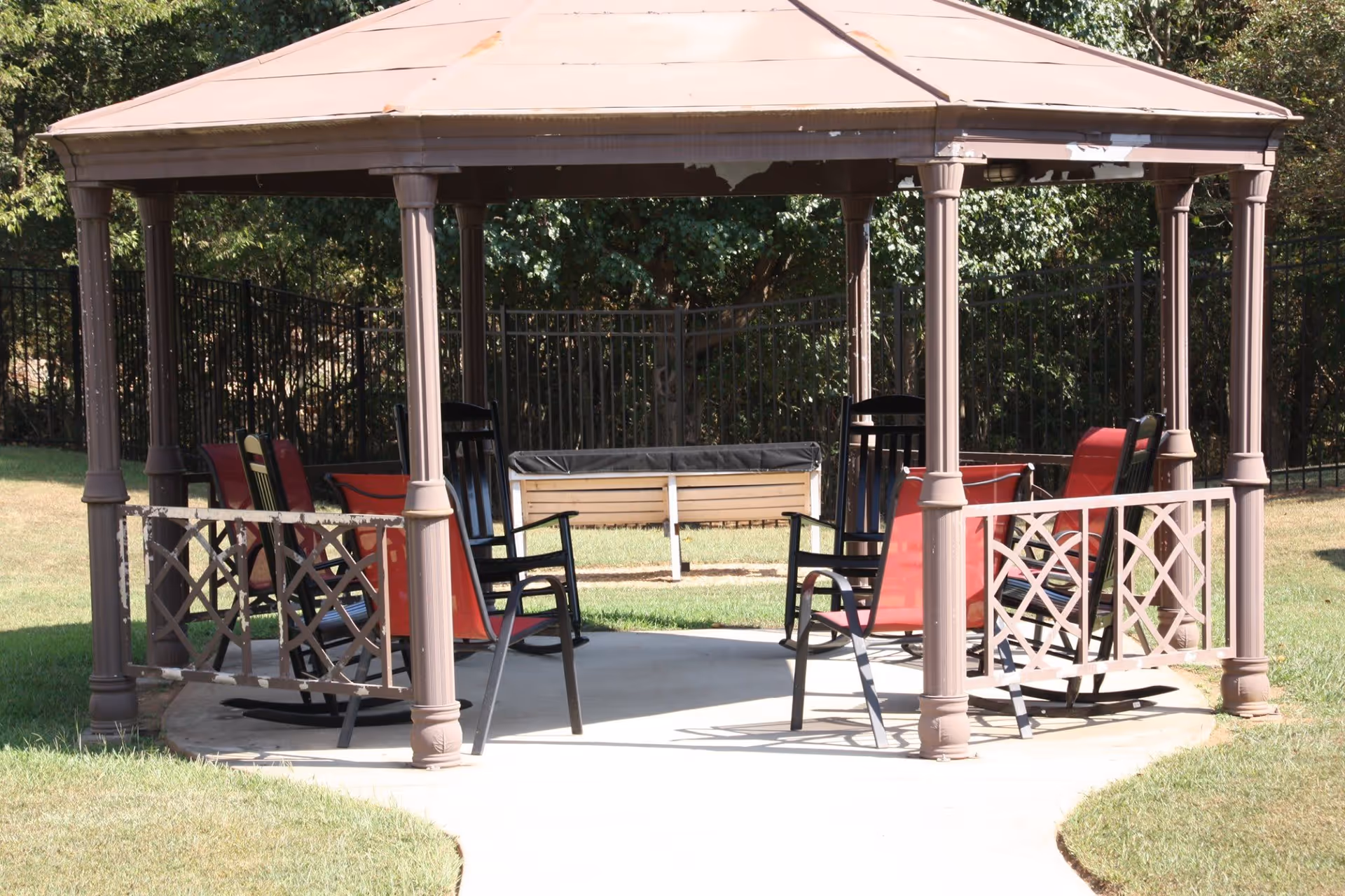 Outdoor gazebo with a brown roof and six chairs arranged inside, including black rocking chairs and red sling chairs, situated on a concrete pad surrounded by grass and trees in the background.