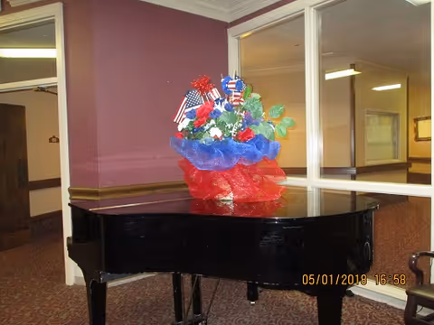 A black grand piano in a carpeted interior space with a patriotic floral arrangement on top, featuring red, white, and blue decorations and small American flags. The walls are painted purple with white trim, and there is a large window looking into another room.