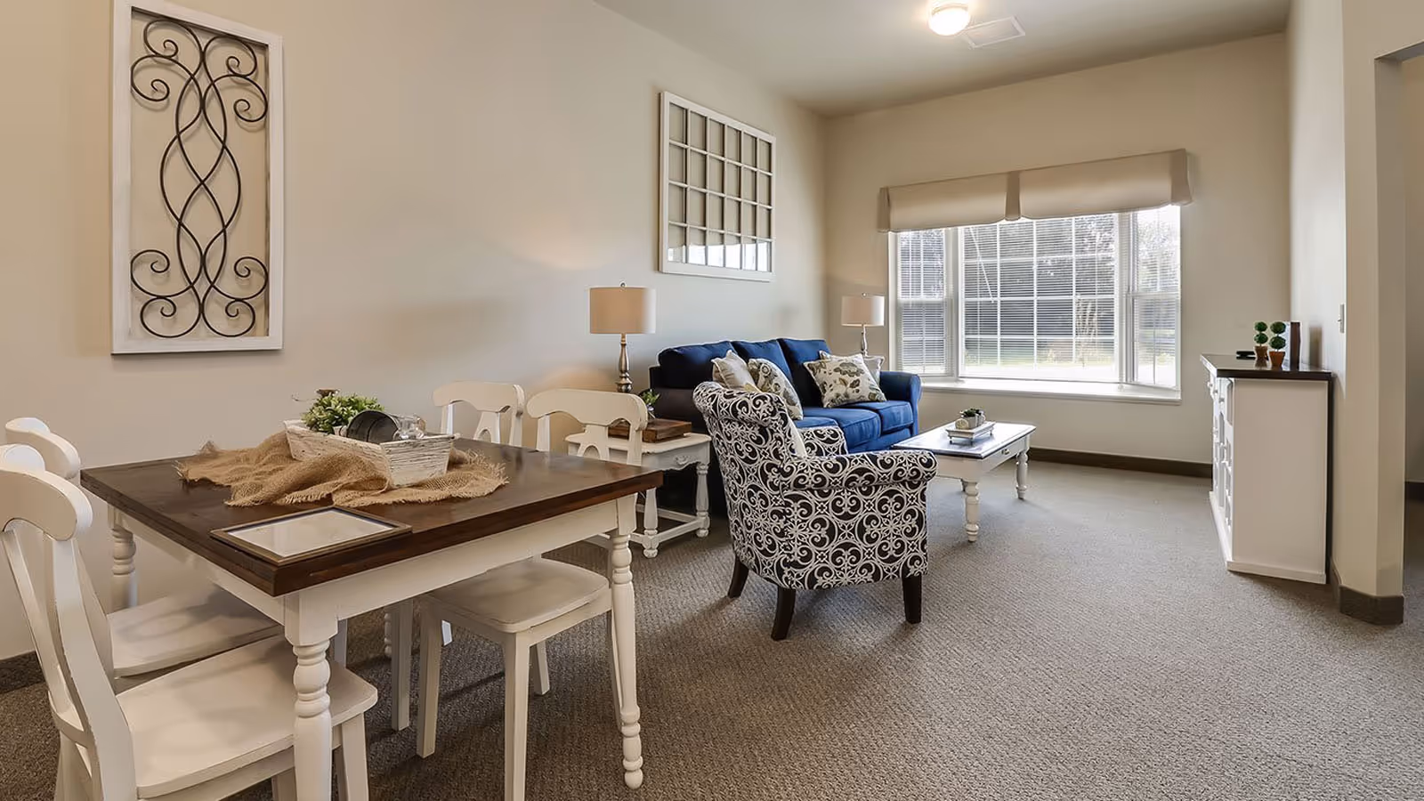 A cozy living room and dining area with a wooden dining table and white chairs on the left, a blue sofa with patterned pillows, a patterned armchair, two table lamps, a white coffee table, and a large window with blinds letting in natural light.