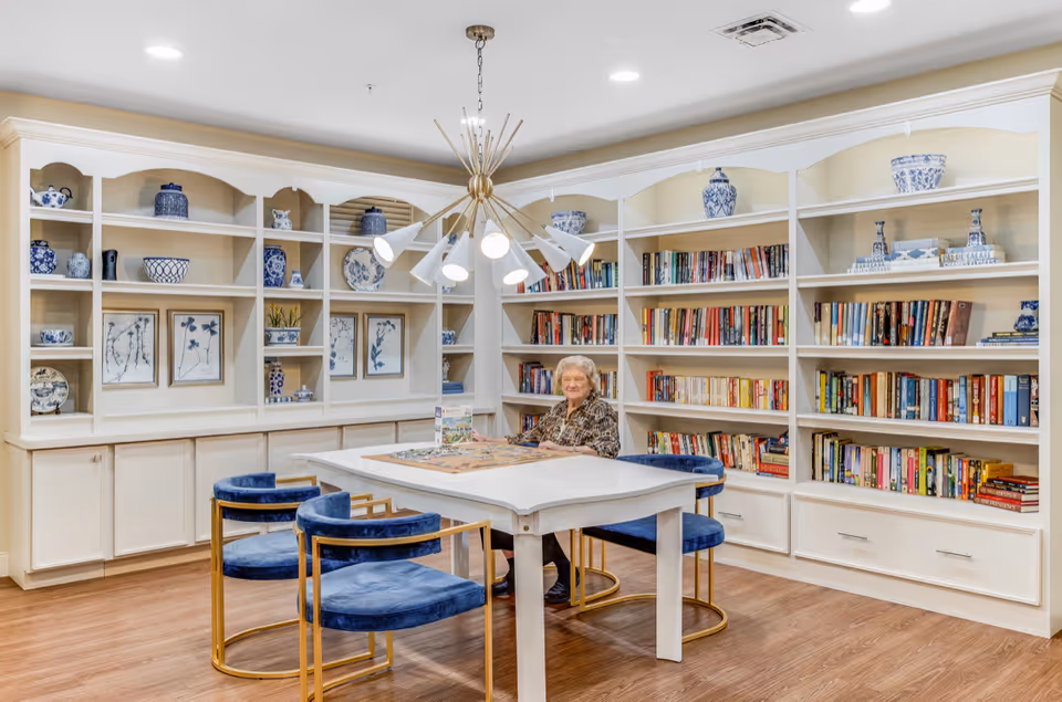 A senior woman sitting at a white table in a cozy library or reading room with built-in white bookshelves filled with books and decorative blue and white ceramics. The room has wooden flooring, blue velvet chairs with gold frames, and a modern chandelier hanging from the ceiling.