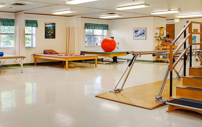 A spacious rehabilitation room with exercise equipment including parallel bars, therapy tables, a large red exercise ball, and a set of stairs with handrails. The room has large windows with green valances, light-colored walls, and a polished floor.