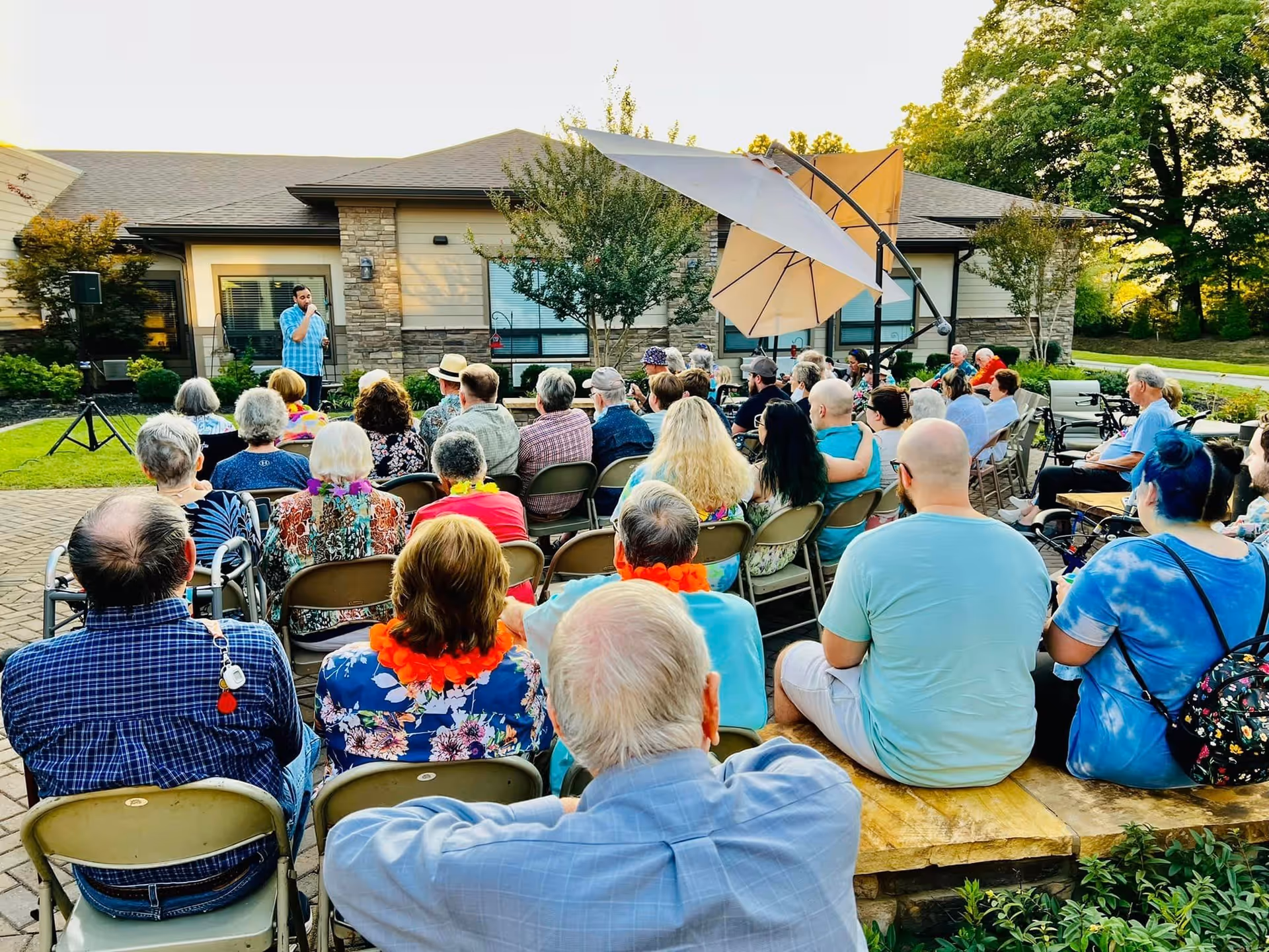 A group of elderly people and some younger adults seated outdoors facing a man speaking into a microphone in front of a senior living facility building. The setting includes chairs arranged in rows, a large umbrella providing shade, and greenery around the paved area.