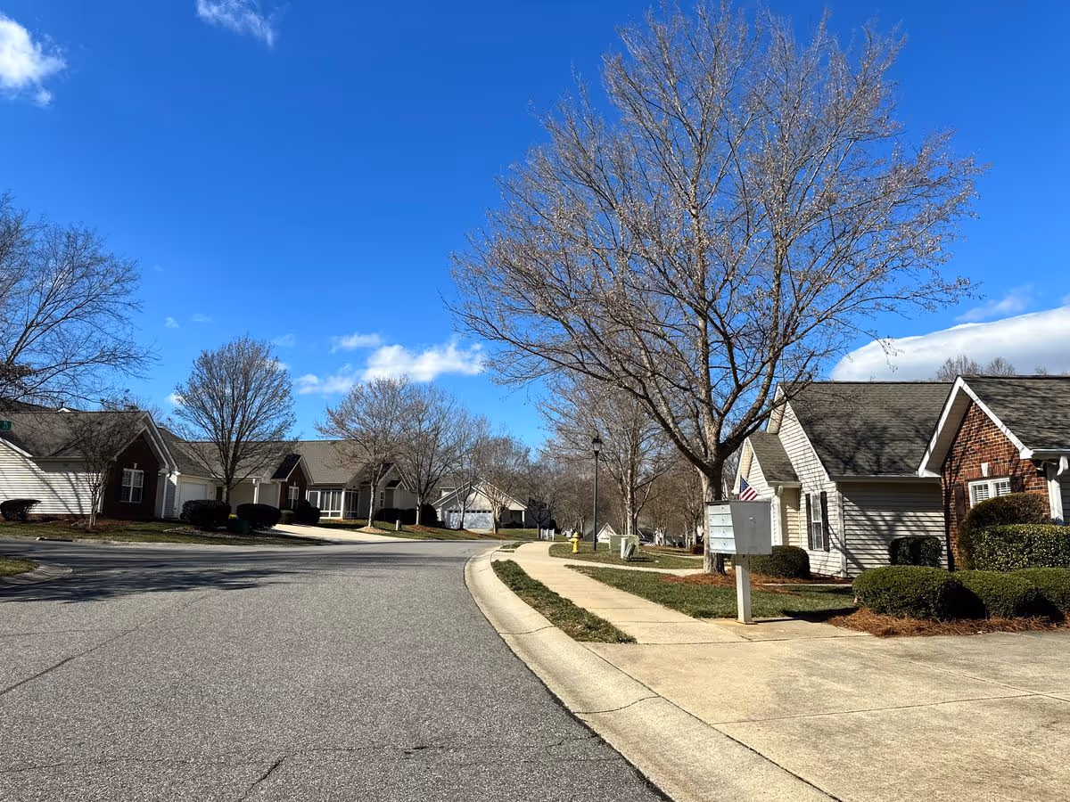 Street view of single-story houses and bare trees along a curving sidewalk under a blue sky.