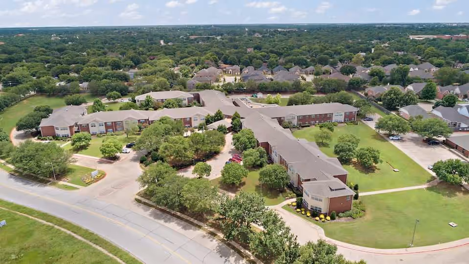 Aerial view of Morada Lake Arlington senior living facility showing multiple connected buildings surrounded by green lawns, trees, and parking areas under a partly cloudy sky.